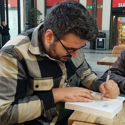 Fotografía de Javer Escámez, un hombre moreno, de pelo corto, barba y gafas, firmando un ejemplar de «Moradores de Covenant». Lleva una sobrecamisa a cuadros grises.